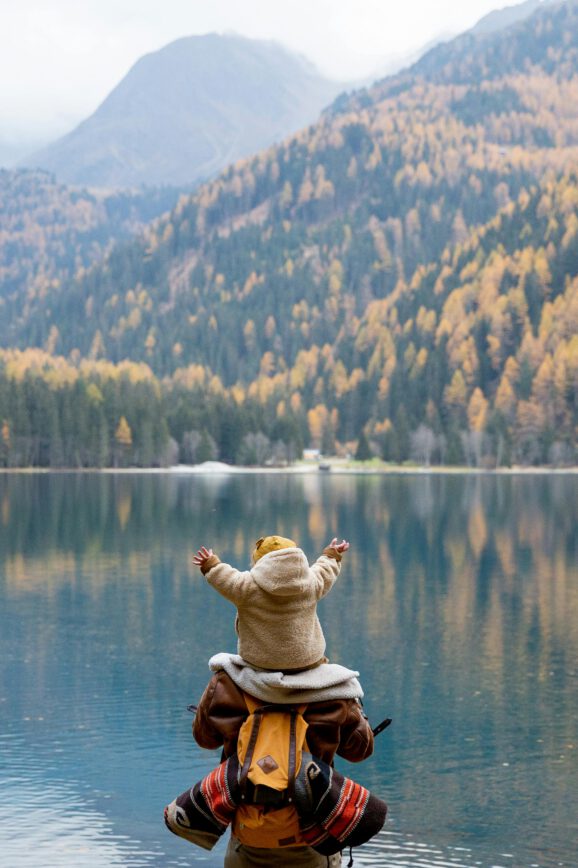 Ein Kind auf den Schultern eines Erwachsenen vor einem Bergsee.
